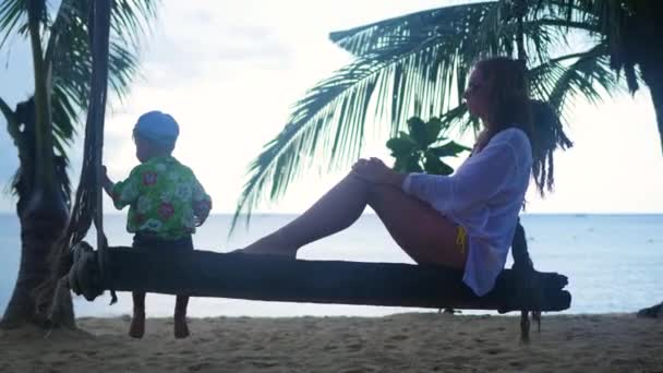 Una niña y un niño balanceándose en un columpio de cuerda en la playa — Vídeos de Stock