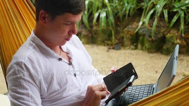 Молодой парень, работающий на ноутбуке и смартфоне, лежащий в гамаке — стоковое видео