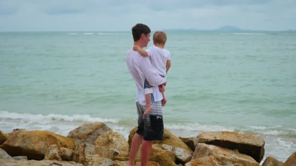 Un joven sosteniendo a un niño en la orilla del mar. Familia feliz — Vídeos de Stock