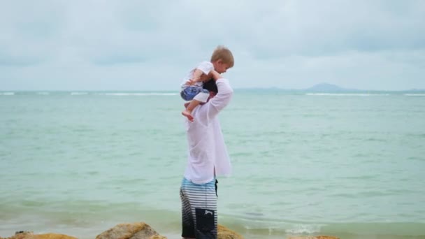 Un joven sosteniendo a un niño en la orilla del mar. Familia feliz — Vídeo de stock
