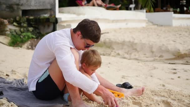 Молодой человек играет с ребенком на пляже. Сделать лепёшки из песка — стоковое видео