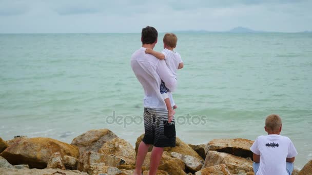 Молодой человек держит ребенка на берегу моря. Счастливая семья — стоковое видео