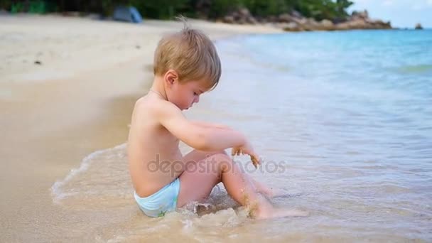 L'enfant est assis sur la plage, nage dans les vagues de la mer par une journée ensoleillée . — Video