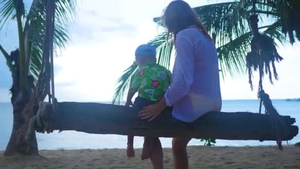 Egy lány és egy gyermek, egy kötélen himbálózott swing a strandon. Trópusi sziget — Stock videók