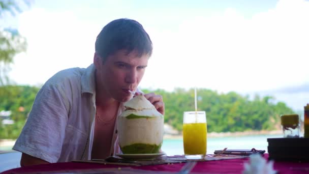 Un joven en un café bebiendo un cóctel en la terraza. al aire libre. Agua de coco — Vídeos de Stock