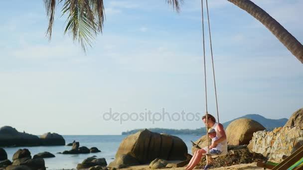 Una niña y un niño balanceándose en un columpio de cuerda en la playa — Vídeos de Stock