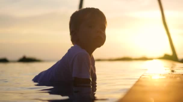 El niño se sienta en el agua y juega con un spray de agua. Tiempo de puesta del sol — Vídeos de Stock