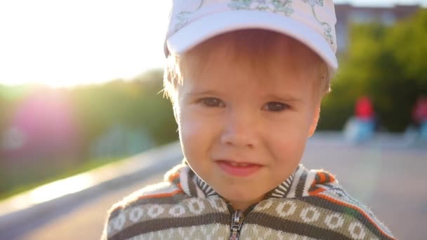 Hermoso niño mira a la cámara y sonríe contra el fondo de los rayos del sol. Diversión al aire libre — Vídeo de stock