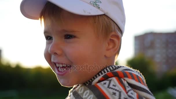 Hermoso niño mira a la cámara y sonríe contra el fondo de los rayos del sol. Diversión al aire libre — Vídeo de stock
