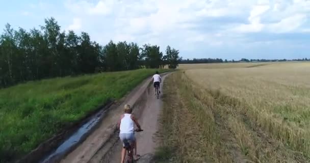 Ein junger Mann mit einem Kind, das auf einer Landstraße entlang der Weizenfelder Fahrrad fährt. Schüsse aus einer Drohne. Sport im Freien — Stockvideo