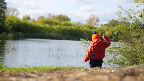 Ένα παιδί στέκεται στην όχθη της λίμνης και πετώντας πέτρες. Περίπατοι στον καθαρό αέρα — Αρχείο Βίντεο