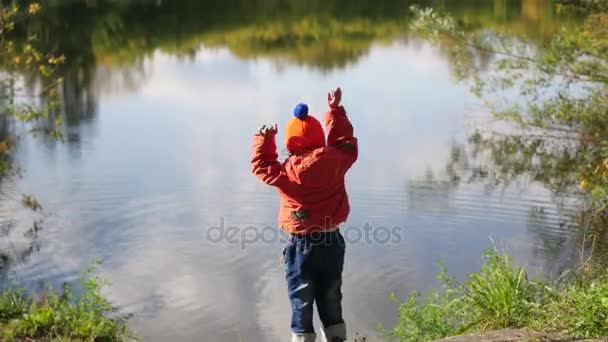 Un niño se para en la orilla del estanque y tira piedras. Caminatas al aire libre — Vídeos de Stock