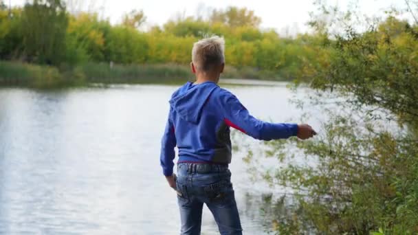 Ένα παιδί στέκεται στην όχθη της λίμνης και πετώντας πέτρες. Περίπατοι στον καθαρό αέρα — Αρχείο Βίντεο