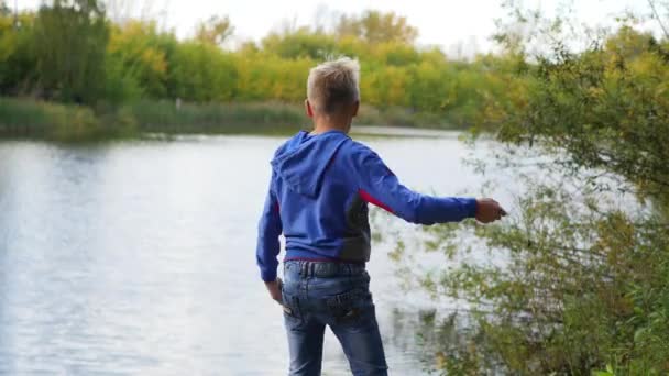 Un niño se para en la orilla del estanque y tira piedras. Caminatas al aire libre — Vídeo de stock