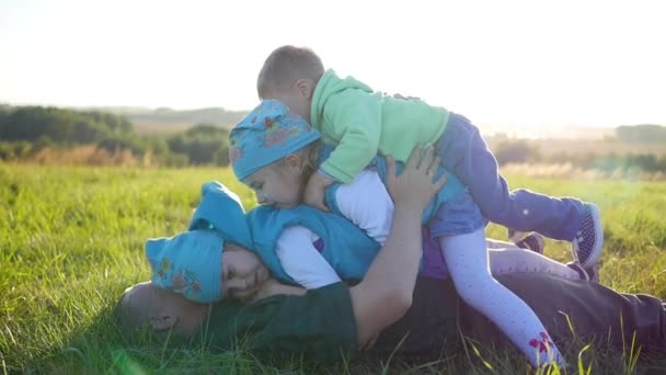 Feliz padre joven con sus hijos se encuentra en el césped en el parque. Tres niños felices juegan con su padre. Vacaciones para niños al aire libre . — Vídeo de stock