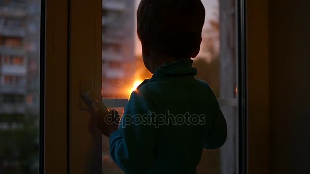 Un niño se para en el alféizar de la ventana y mira por la ventana hacia el sol. Puesta de sol — Vídeos de Stock