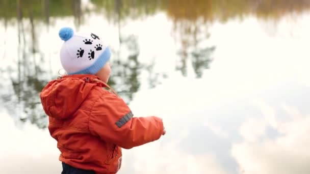 Un niño se para en la orilla del estanque y tira piedras. Caminatas al aire libre — Vídeos de Stock