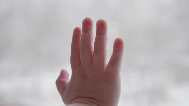 Un bambino sta vicino a una finestra e guarda la neve cadere per strada. La mano di primo piano sulla finestra di vetro — Video Stock