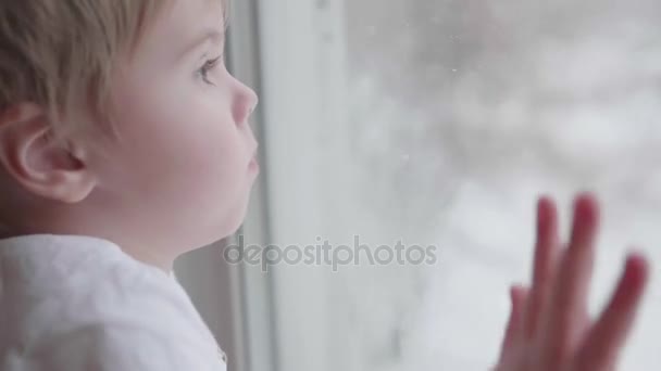 Un bambino sta vicino a una finestra e guarda la neve cadere per strada. La mano di primo piano sulla finestra di vetro — Video Stock