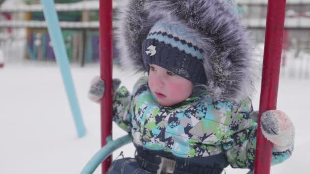 Een klein kind speelt in winter Park. Een kind op een schommel. Een zonnige winterdag. Pret en spelen in de frisse lucht. — Stockvideo