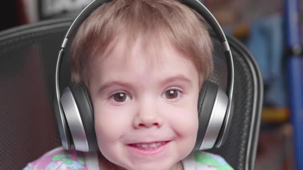 Un niño gracioso se sienta en una silla y escucha música a través de auriculares. Cara de cerca — Vídeo de stock