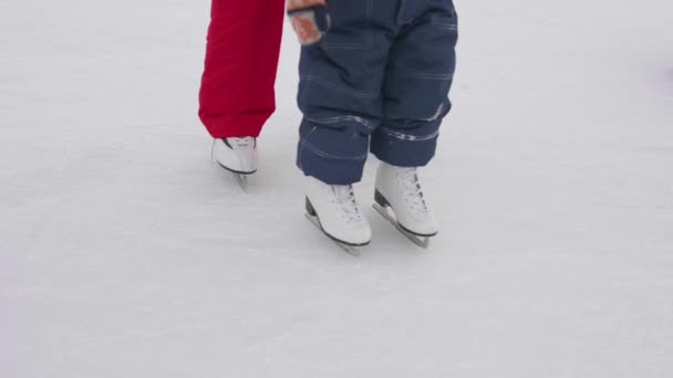 Первые шаги маленького ребенка на коньках. Мама учит кататься на коньках и держит его ребенка на льду. Активный образ жизни — стоковое видео