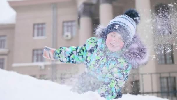 Ett litet barn spelar på en snöig berg, kastar snö och skrattar. Solig frostig dag. Kul och spel i friska luften. — Stockvideo