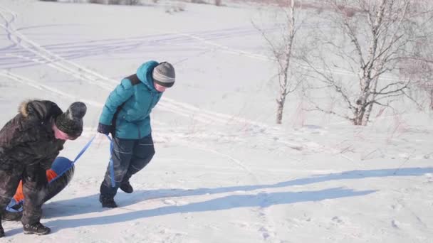 Duas crianças montam em uma colina nevada em um trenó. As crianças sobem ao topo da montanha. Caminhadas ao ar livre — Vídeo de Stock