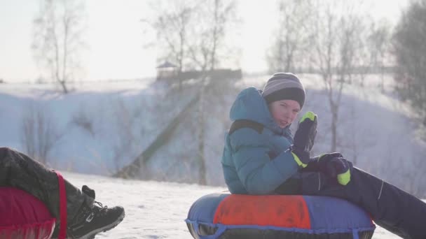 Dos niños cabalgan en una colina nevada en un trineo. Los niños se sientan en la cima de la montaña. Deportes y actividades al aire libre — Vídeo de stock