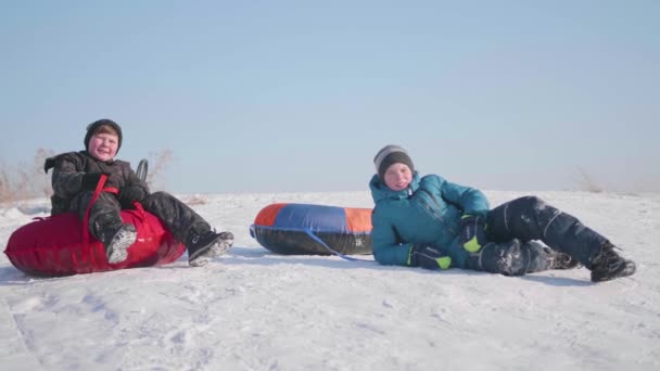 Dos niños cabalgan en una colina nevada en un trineo. Los niños se sientan en la cima de la montaña. Deportes y actividades al aire libre — Vídeos de Stock