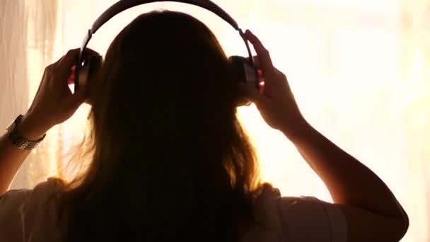 Молода дівчина приходить до вікна і одягає навушники, щоб слухати музику. Розмитий фон із заходом сонця, жінка насолоджується мелодією в навушниках . — стокове відео