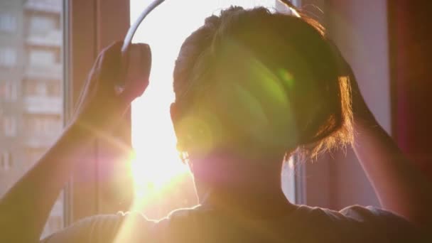 Uma jovem vem à janela e veste fones de ouvido para ouvir música. Fundo borrado com pôr do sol, mulher desfrutando da melodia em fones de ouvido . — Vídeo de Stock