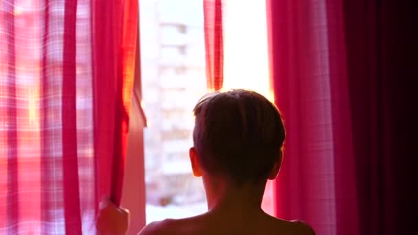 Killen kommer tidigt på morgonen till fönstret, öppnar gardiner. Solens strålar passerar genom glaset. — Stockvideo