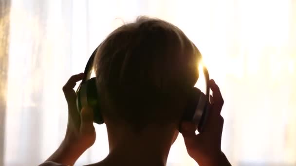 Genç bir adam pencerenin yanında duruyor ve müzik dinlemek için kulaklık takıyor. Bulanık arka plan, günbatımı, gençlerin kulaklıkla müzik dinlemesi.. — Stok video