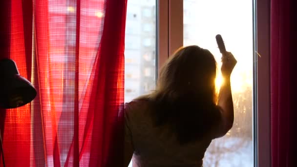 Chica temprano en la mañana de pie en la ventana y se peina el pelo. Los rayos del sol pasan a través del cristal iluminan la habitación y la chica con luz de la mañana — Vídeos de Stock