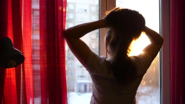 Menina no início da manhã em pé na janela e penteia o cabelo. Os raios de sóis passam pelo vidro iluminam o quarto e a menina com luz da manhã — Vídeo de Stock