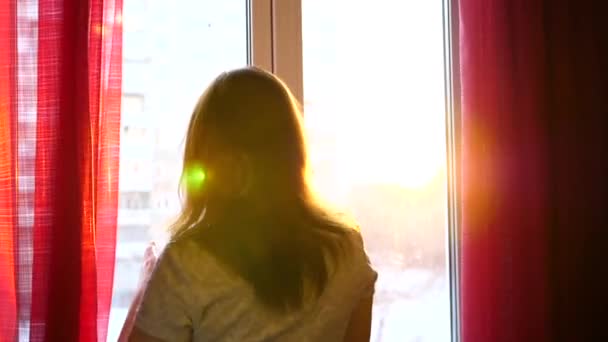 Una niña llega a la ventana y viste auriculares para escuchar música. Fondo borroso con puesta de sol, mujer disfrutando de la melodía en auriculares. Los rayos del sol pasan a través del vidrio — Vídeo de stock