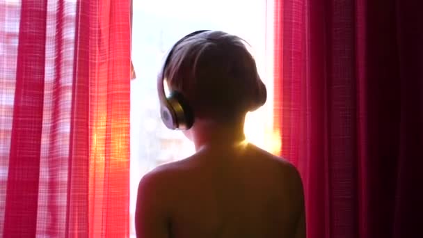 El joven se para junto a la ventana y baila, escuchando música a través de auriculares. Fondo borroso con puesta de sol, chico disfrutando de la música en los auriculares . — Vídeo de stock