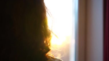 Yakın çekim. Genç bir kız pencereye gelir ve müzik dinlemek için kulaklık elbiseler. Gün batımı, kadının Kulaklığımda melodi ile arka plan bulanık. Güneş ışınları camı delip geçmek