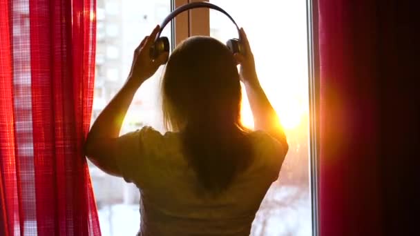 Молода дівчина приходить до вікна і одягає навушники, щоб слухати музику. Розмитий фон із заходом сонця, жінка насолоджується мелодією в навушниках. Промені сонця проходять через скло — стокове відео