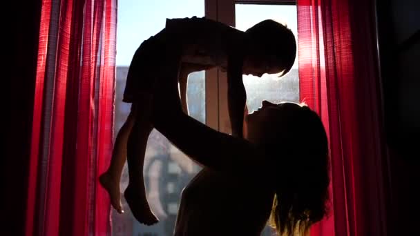 A young and happy mother is holding her child next to the window. The suns rays pass through the window. The laughter and joy of a child and mother. — Stock Video