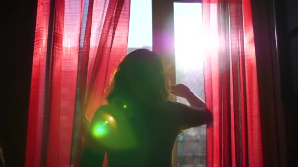 La chica de madrugada camina hacia la ventana y la abre. Los rayos del sol pasan a través del cristal, iluminan la habitación y la niña con luz de la mañana — Vídeos de Stock