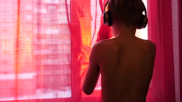 El joven se acerca a la ventana, usa auriculares para escuchar música. Fondo borroso con puesta de sol, adolescente disfrutando de la música en los auriculares . — Vídeo de stock