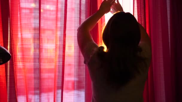 Jovencita feliz viniendo a la ventana, abre las cortinas. Los rayos del sol pasan a través de la ventana, iluminando la habitación y la niña. Tiempo de puesta del sol — Vídeos de Stock