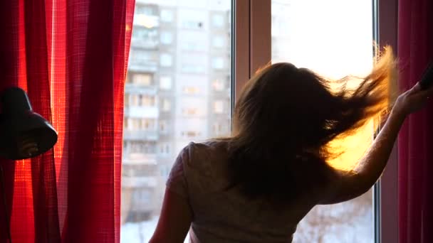 Девочка рано утром стоит у окна и расчесывает волосы. Солнечные лучи проходят через стекло освещая комнату и девушку утренним светом — стоковое видео