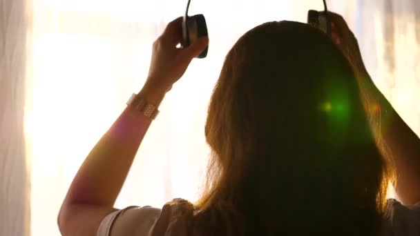 Una joven llega a la ventana y viste auriculares para escuchar música.Mujer disfrutando de la música en los auriculares . — Vídeos de Stock