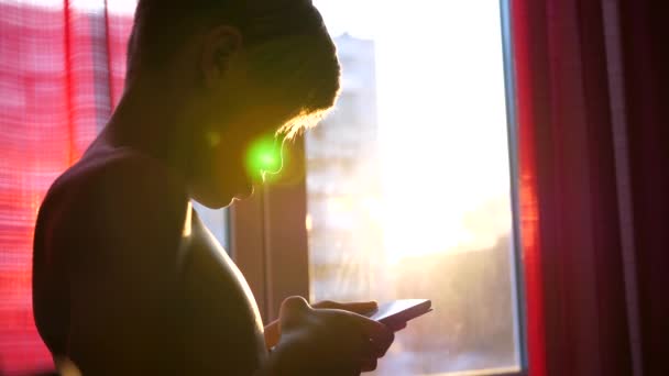 Молодой парень стоит у окна с телефоном в руках. Время заката, солнечные лучи проходят в комнату через стекло — стоковое видео