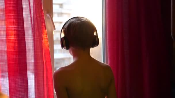 Den unge mannen står vid fönstret och danser, lyssna på musik i hörlurar. Killen har musiken i hörlurarna. — Stockvideo