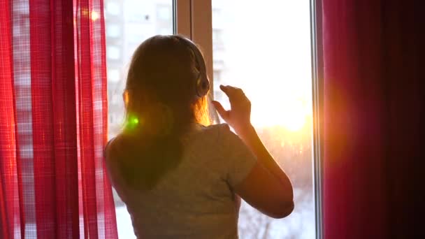 Молода дівчина приходить до вікна і одягає навушники, щоб слухати музику. Дівчина насолоджується музикою з навушниками, танцями. Промені сонця проходять через скло — стокове відео
