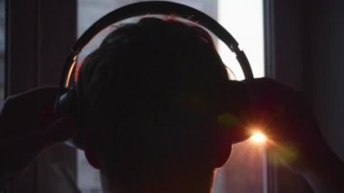Yakın çekim. Genç bir adam pencerenin duruyor ve müzik dinlemek için kulaklık koyar. Gün batımı ile arka plan bulanık adam zevk müzik kulaklık.